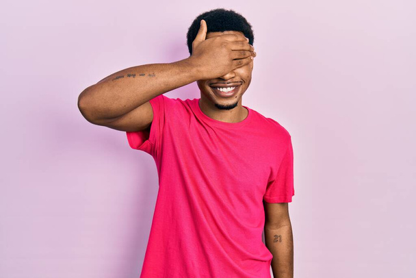 Young african american man wearing casua t shirt smiling and laughing with hand on face covering eyes for surprise. blind concept.  - Photo, image