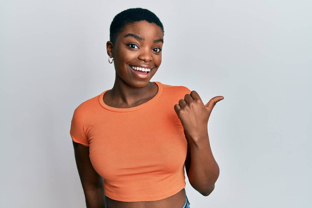 Νεαρή Αφροαμερικανή που φοράει πορτοκαλί μπλουζάκι χαμογελώντας με χαρούμενο πρόσωπο και δείχνοντας στο πλάι με τον αντίχειρα προς τα πάνω.  - Φωτογραφία, εικόνα
