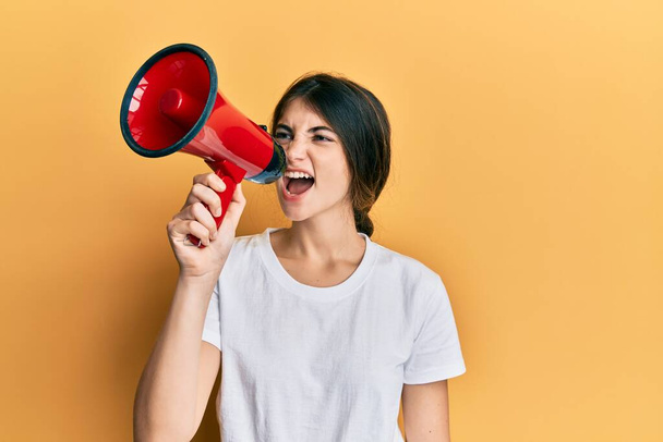 young beautiful woman holding megaphone over isolated yellow background surprised and shocked shouting to the side - Foto, Bild