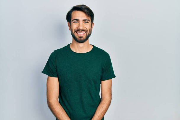 Fiatal spanyol férfi alkalmi fehér pólóban, boldog és hűvös mosollyal az arcán. szerencsés ember.  - Fotó, kép