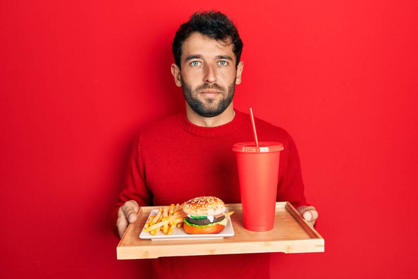 Handsome man with beard eating a tasty classic burger with fries and soda relaxed with serious expression on face. simple and natural looking at the camera.  - Foto, imagen