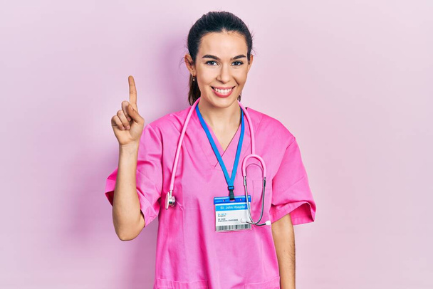 Νεαρή μελαχρινή γυναίκα με στολή γιατρού και στηθοσκόπιο που δείχνει και δείχνει προς τα πάνω με το δάχτυλο νούμερο ένα, ενώ χαμογελά αυτοπεποίθηση και χαρούμενος.  - Φωτογραφία, εικόνα