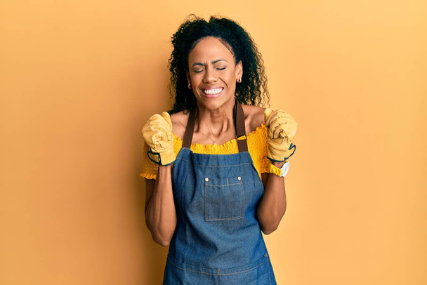 Middle age african american woman wearing professional apron excited for success with arms raised and eyes closed celebrating victory smiling. winner concept.  - Foto, Bild