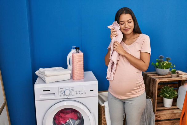Молода латинка вагітна тримає дитячий одяг у пральні
 - Фото, зображення
