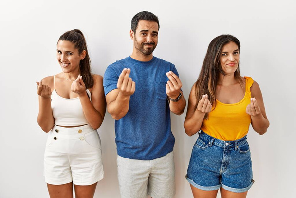 Grupo de jóvenes hispanos de pie sobre aislados antecedentes haciendo gesto de dinero con las manos, pidiendo pago de sueldo, negocio millonario  - Foto, imagen