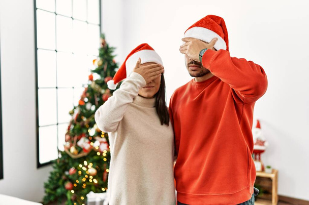 Genç İspanyol çift Noel ağacının yanında dikiliyor. Elleriyle gözlerini kapatıyor. Ciddi ve üzgün görünüyorlar. Görme, saklama ve reddetme konsepti  - Fotoğraf, Görsel