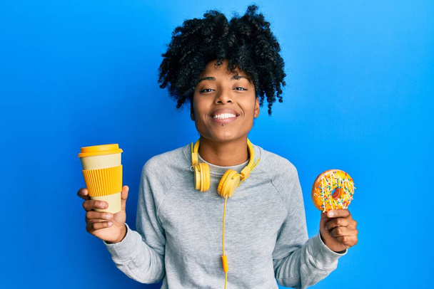 Mujer afroamericana con el pelo afro comiendo donut y bebiendo café sonriendo con una sonrisa alegre y fresca en la cara. mostrando dientes.  - Foto, Imagen