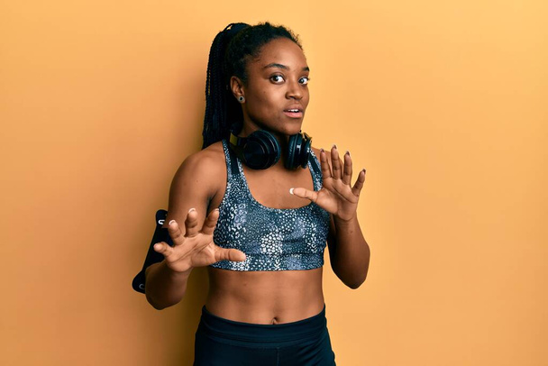 African american woman with braided hair wearing sportswear and arm band afraid and terrified with fear expression stop gesture with hands, shouting in shock. panic concept.  - Foto, afbeelding