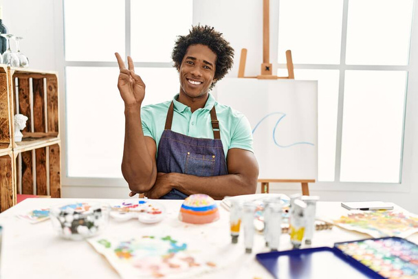 Mladý africký Američan, sedící na stole v ateliéru umění a usmívající se s veselou tváří, mrkající na kameru a předvádějící znamení vítězství. číslo dvě.  - Fotografie, Obrázek