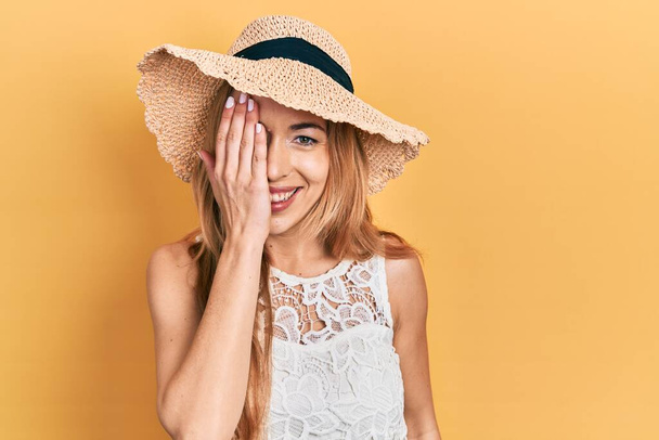 Młoda kaukaska kobieta w letniej czapce zakrywającej jedno oko ręką, pewny siebie uśmiech na twarzy i niespodziewane emocje.  - Zdjęcie, obraz