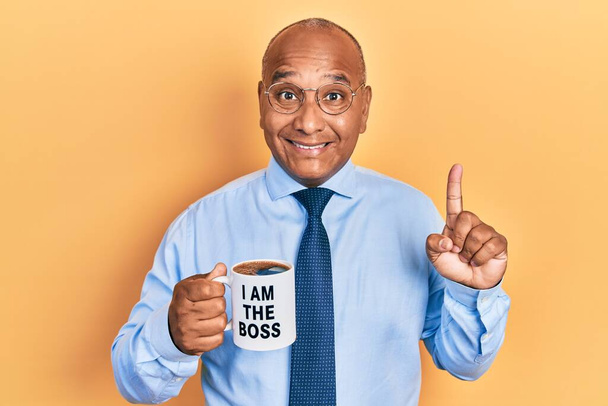 Middle age latin man drinking from i am the boss coffee cup smiling with an idea or question pointing finger with happy face, number one  - Foto, imagen