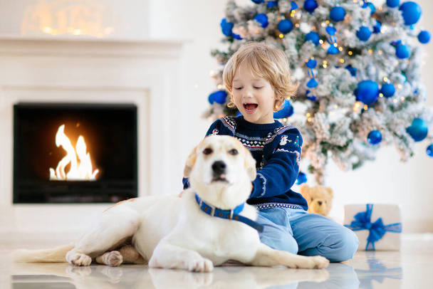 Christmas at home. Kids and dog under Xmas tree. Little boy hug pet in Santa hat and open Christmas presents. Children play with animal. Winter holiday celebration. Blue and white theme. - Photo, image