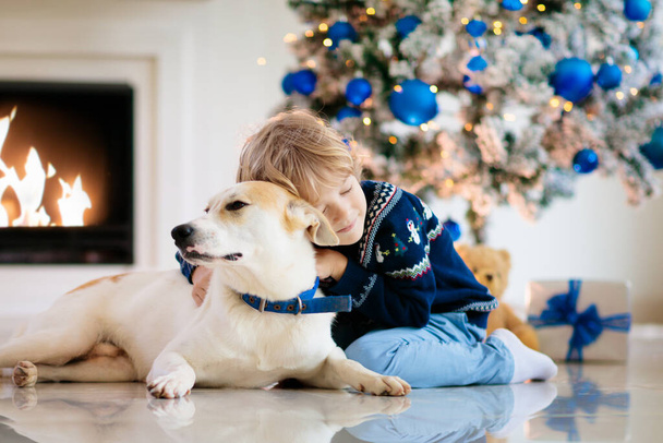 Joulu kotona. Lapset ja koira joulukuusen alla. Pieni poika halata lemmikki Santa hattu ja avaa joululahjoja. Lapset leikkivät eläimillä. Talviloma juhla. Sininen ja valkoinen teema. - Valokuva, kuva