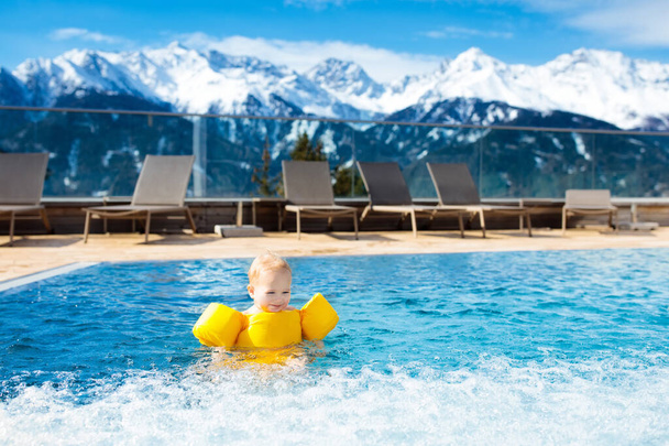 Маленький ребенок играет в открытом бассейне роскошного альпийского курорта в горах Альп, Австрия. Зимний и снежный отдых с детьми. джакузи на открытом воздухе с видом на горы. Дети играют и плавают
. - Фото, изображение