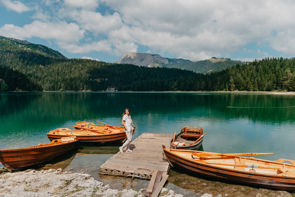 The girl looks at the Black Lake with fishing boats, forest and mountains in the Durmitor National Park of Montenegro. A tourist enjoys a magical view of the lake, a coniferous forest and a magical - Fotoğraf, Görsel