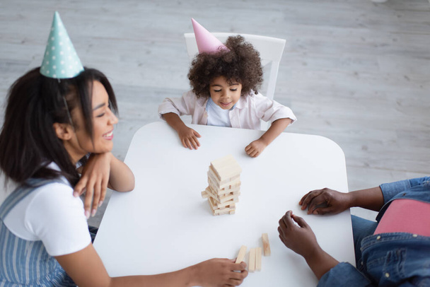 hoge hoek uitzicht op Afrikaans amerikaans kind in party cap spelen houten blokken spel met glimlachende moeder en oma - Foto, afbeelding