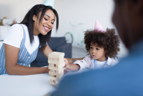 smiling african american woman and girl in party caps playing wood blocks game near blurred granny - Φωτογραφία, εικόνα