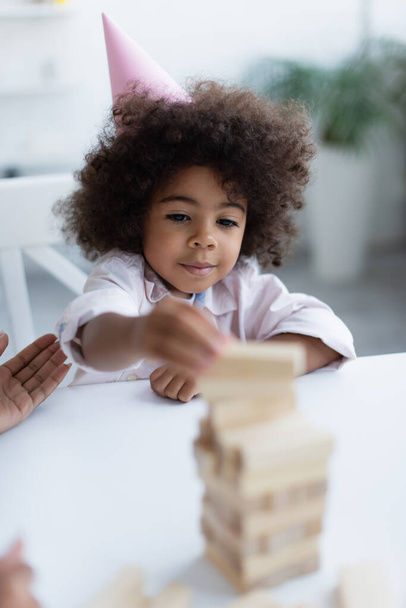 encaracolado criança afro-americana em boné de festa jogando jogo de blocos de madeira em primeiro plano borrado - Foto, Imagem