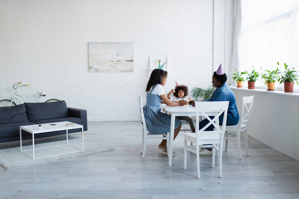 african american women and girl in party caps playing wood blocks game in spacious living room - Foto, Bild