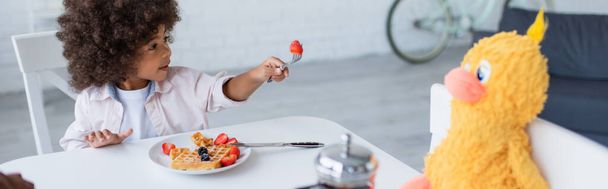 アフリカ系アメリカ人の子供が台所で朝食中におもちゃの雛にイチゴを提案 - 写真・画像