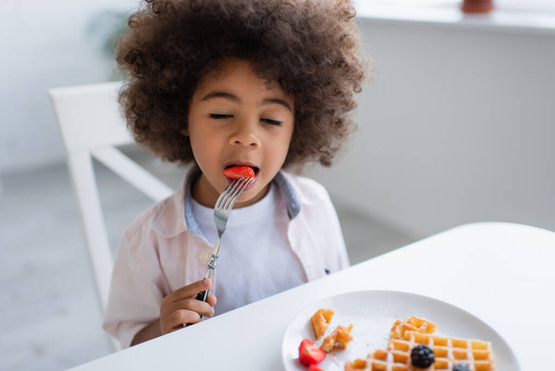 curly african american girl with closed eyes eating strawberry near tasty waffle on plate - Фото, изображение