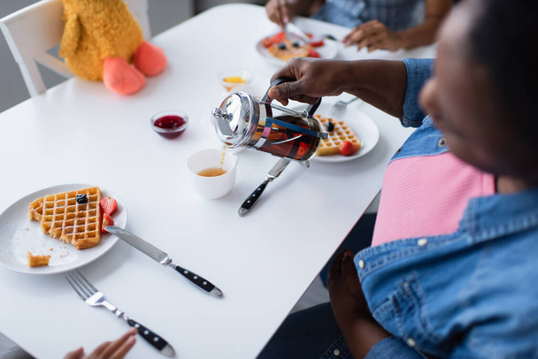 high angle view of blurred african american woman pouring tea near tasty waffles with berries during breakfast with family - Photo, Image