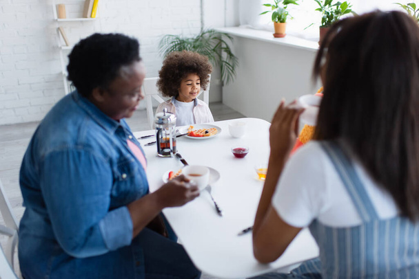 mujeres afroamericanas borrosas tomando té durante el desayuno con una niña en la cocina - Foto, imagen