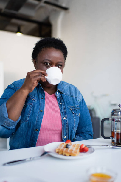 mujer afroamericana de mediana edad bebiendo té cerca de gofre borroso con bayas frescas en el plato - Foto, imagen