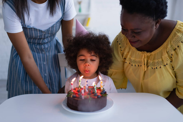 アフリカ系アメリカ人の女の子がおばあちゃんとお母さんの近くの誕生日ケーキにろうそくを吹き - 写真・画像