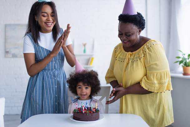wesoły Afryki amerykanki klaskanie ręce w pobliżu dziewczynka maluch i tort urodzinowy - Zdjęcie, obraz