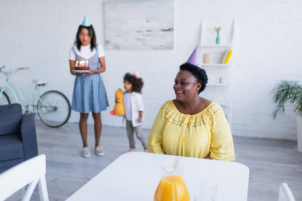happy mature african american woman in party cap near blurred family with birthday cake - Photo, Image