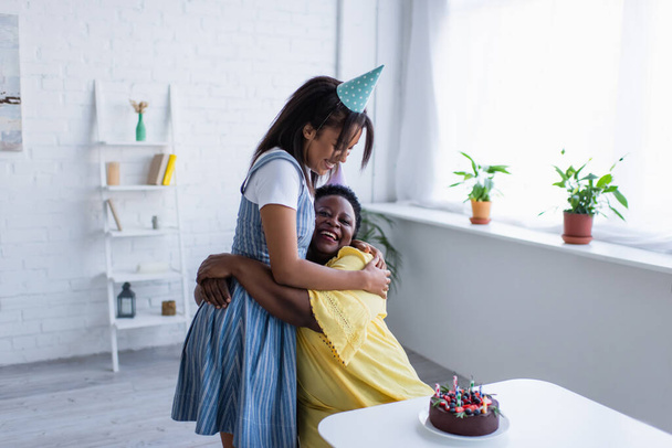 giovane donna afroamericana in berretto da festa che abbraccia la madre felice vicino alla torta di compleanno con le candele   - Foto, immagini
