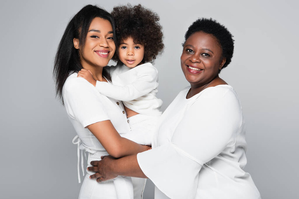 compiaciute donne africane americane in abiti bianchi guardando la fotocamera mentre tenevano il bambino isolato sul grigio - Foto, immagini