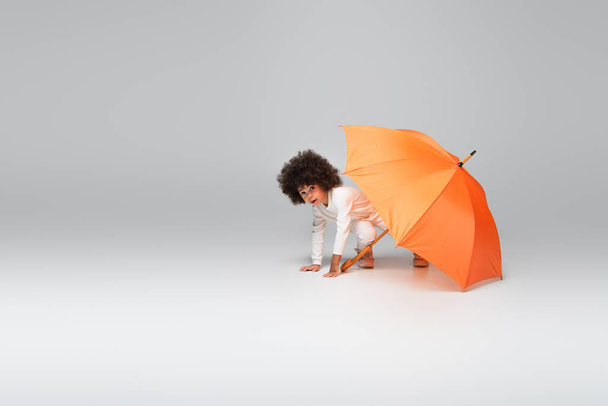 afro-américaine fille en blanc vêtements regardant caméra tout en rampant sous parapluie orange sur gris - Photo, image