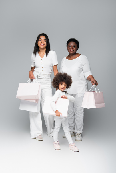 smiling african american kid with mother and granny holding white shopping bags on grey - Photo, image