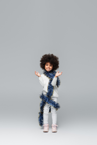 full length view of cheerful african american girl in white clothes posing with christmas tinsel on grey - Photo, Image