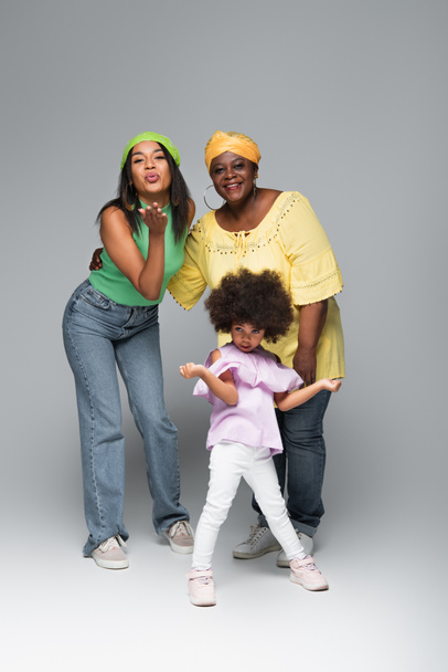 elegante donna afro-americana che soffia aria bacio vicino a madre e figlia sul grigio - Foto, immagini
