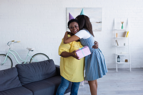 young african american woman in party cap embracing happy mother holding birthday present  - Foto, Imagem