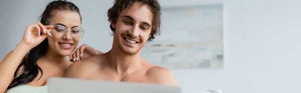 Shirtless man looking at laptop near smiling girlfriend in eyeglasses at home, banner  - Foto, immagini