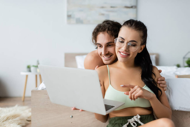 Lächelnde Frau mit Top und Brille, die auf Laptop neben hemdlosem Freund im Schlafzimmer zeigt  - Foto, Bild