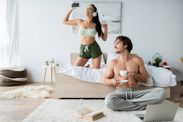 Shirtless man holding cup near laptop, book and smiling girlfriend in headphones in bedroom - Foto, Imagem