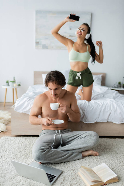 Shirtless freelancer holding cup near laptop and sexy girlfriend in headphones on bed  - Fotografie, Obrázek