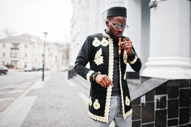 Mega stylish african man in traditional jacket pose. Fashionable black guy in hat and sunglasses with cigar in hand. - Foto, Bild