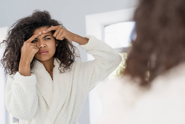 Upset young afro american woman squeeze pimple on forehead, looking at mirror, standing in bathroom. Female in bathrobe worried about acne or skin breaking out pores. Skincare, cosmetology concept - Фото, изображение