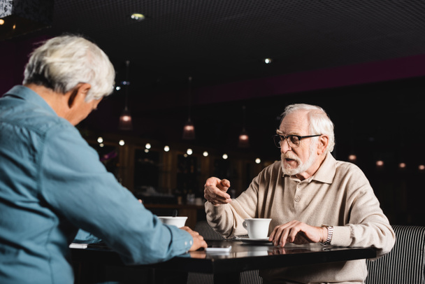 elderly man in eyeglasses talking to blurred friend near cups of coffee - 写真・画像