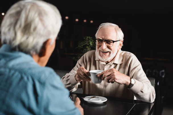joyful senior man in eyeglasses holding coffee cup near blurred friend in bar - Photo, image