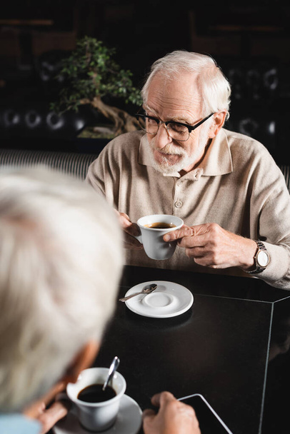 senior muž v brýlích drží šálek kávy v blízkosti rozmazané přítel v kavárně - Fotografie, Obrázek