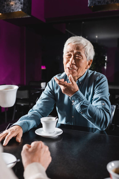 позитивный азиатский пожилой мужчина жестикулирует во время разговора с размытым другом в кафе - Фото, изображение