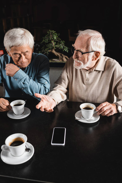 старший человек в очках разговаривает с азиатским другом возле чашек кофе и смартфона с чистым экраном - Фото, изображение