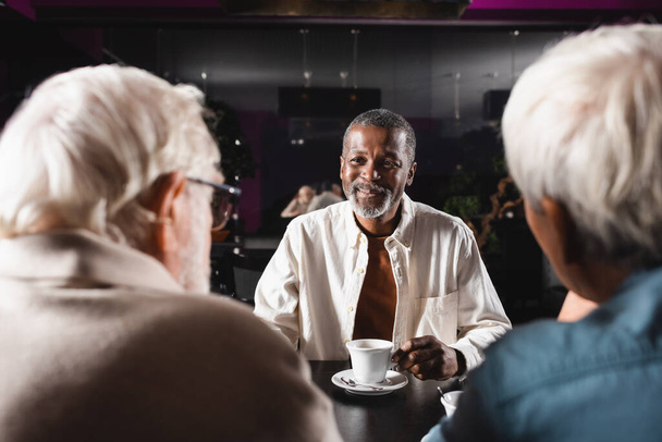 Bir fincan kahvenin yanında oturan mutlu Afrikalı Amerikalı adam ve bulanık kıdemli arkadaşları - Fotoğraf, Görsel